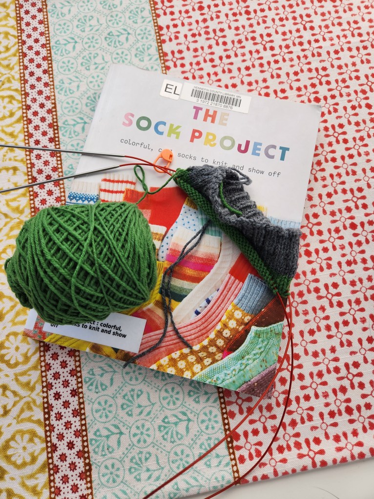 The Sock Project book with a newly cast on sock on top featuring gray yarn for the cuff and green for the leg portion. 