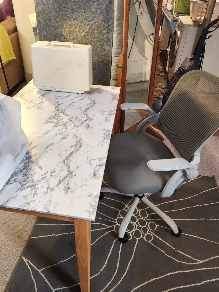 Cozy basement sewing space with functional desk chair, large patterned floral rug in gray and white, and a table covered with white and gray marbled contact paper. 