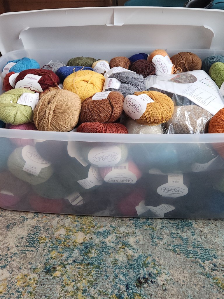 A large, clear plastic storage tub overflowing with various skeins of colorful "Knit Picks" yarn in shades of yellow, blue, green, and terracotta.