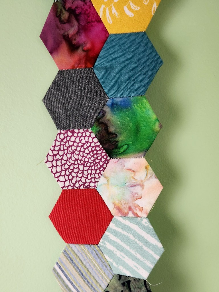 English Paper Piecing rows made from a colorful variety of fabric scraps held against the wall in which the finished project will hang. 